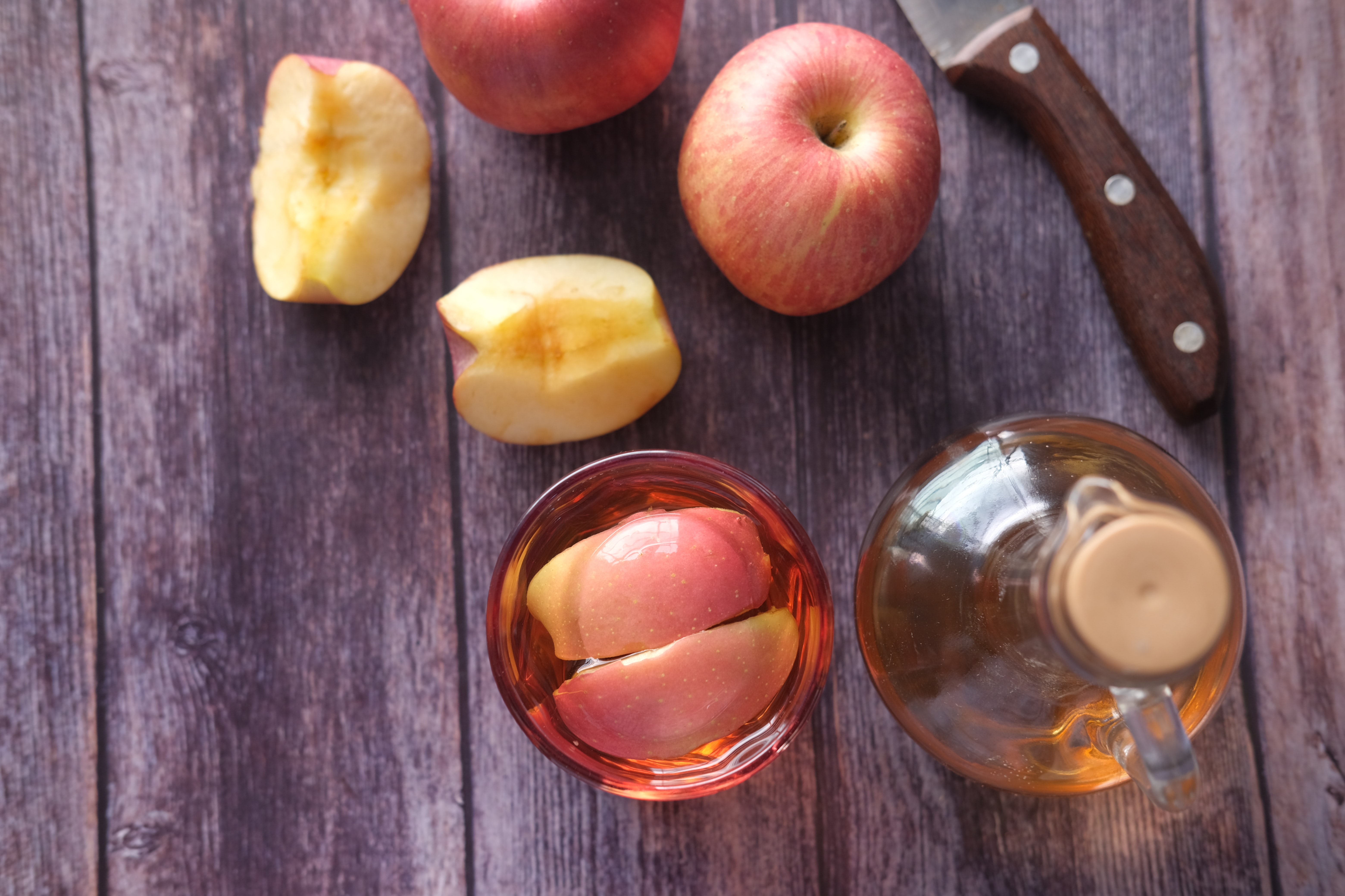 A wood table with various types of apples, one apple is cut and half of it is placed in a cup of apple juice